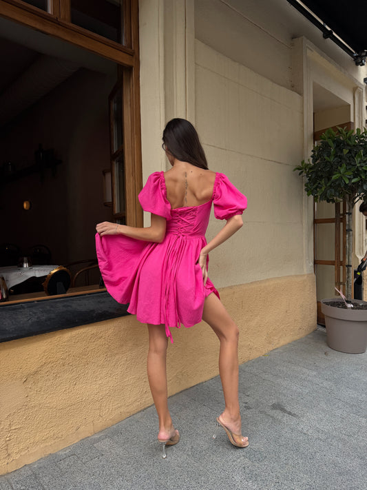 A woman posing from behind wearing a vibrant hot pink off-the-shoulder puff-sleeve mini dress with a tie-back detail, paired with clear high-heeled sandals, standing on a sidewalk in front of a beige building.