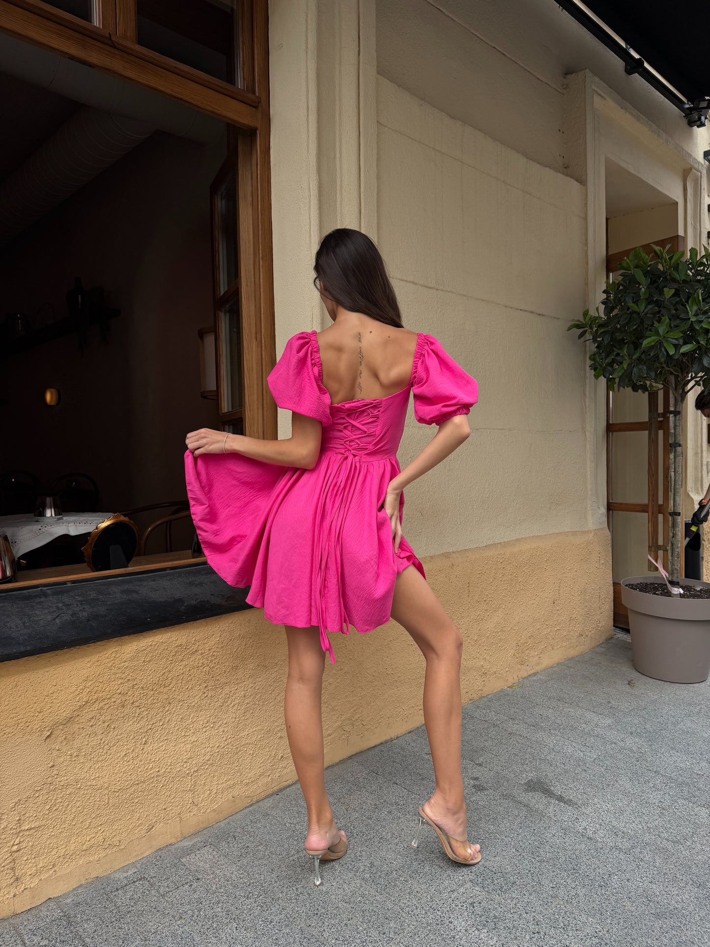 A woman posing from behind wearing a vibrant hot pink off-the-shoulder puff-sleeve mini dress with a tie-back detail, paired with clear high-heeled sandals, standing on a sidewalk in front of a beige building.
