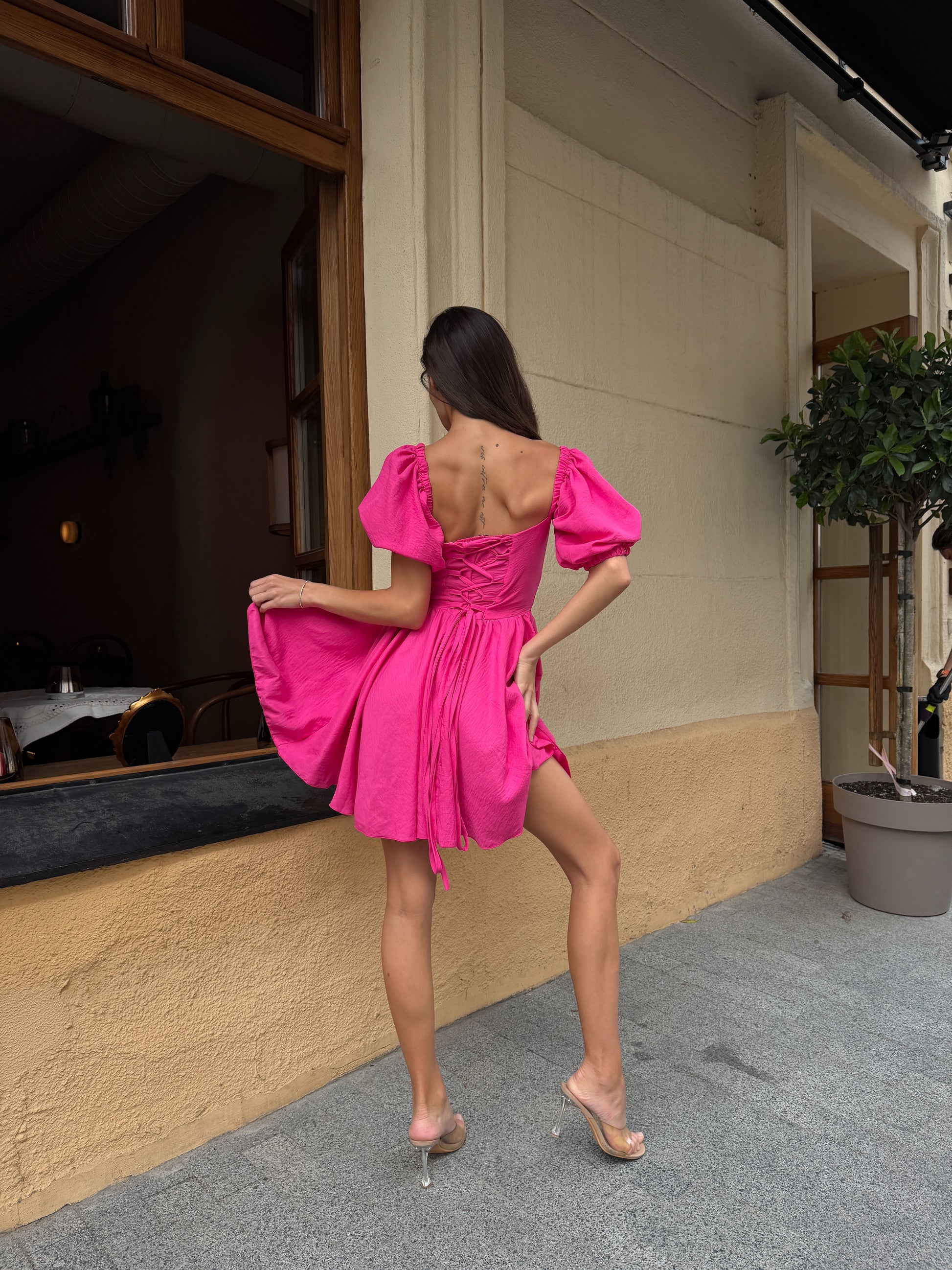 A woman posing from behind wearing a vibrant hot pink off-the-shoulder puff-sleeve mini dress with a tie-back detail, paired with clear high-heeled sandals, standing on a sidewalk in front of a beige building.