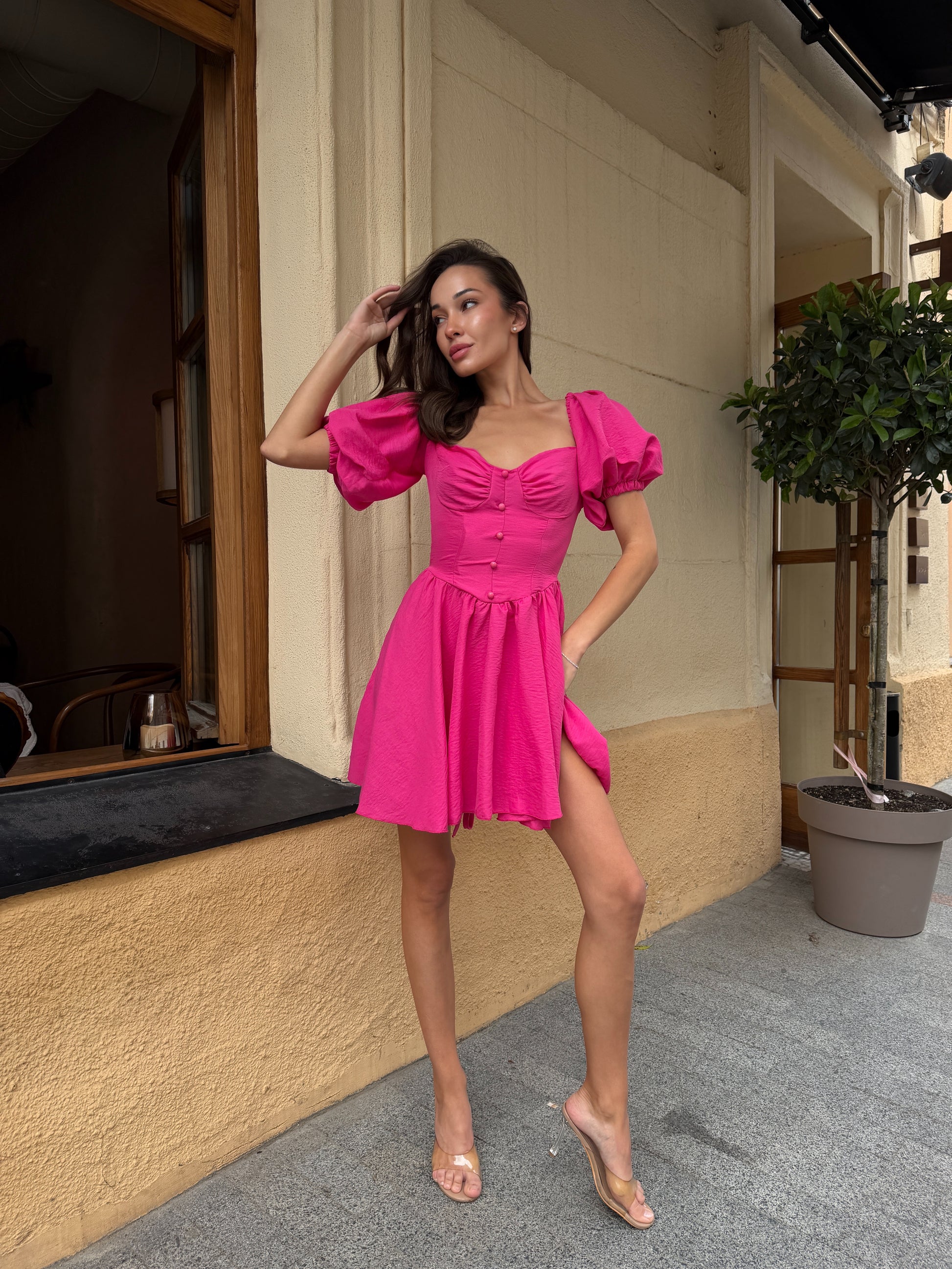A woman models a bright pink puff-sleeve mini dress with a sweetheart neckline and button details, standing on a sidewalk next to a beige building.