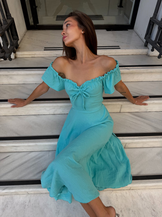 A woman wearing a turquoise off-the-shoulder midi dress sits on white marble steps.