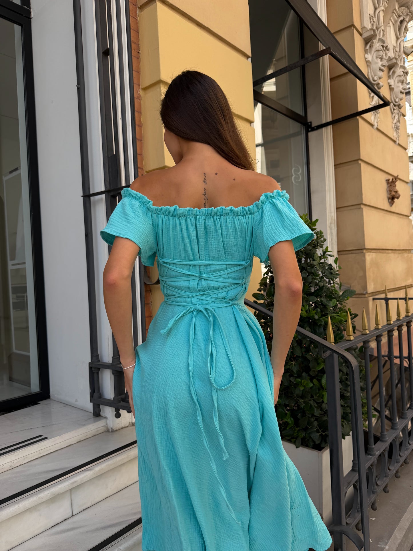 A woman viewed from behind wearing a bright turquoise off-the-shoulder midi dress with a gathered waist and tie detail, standing on a balcony with a beige building facade in the background.
