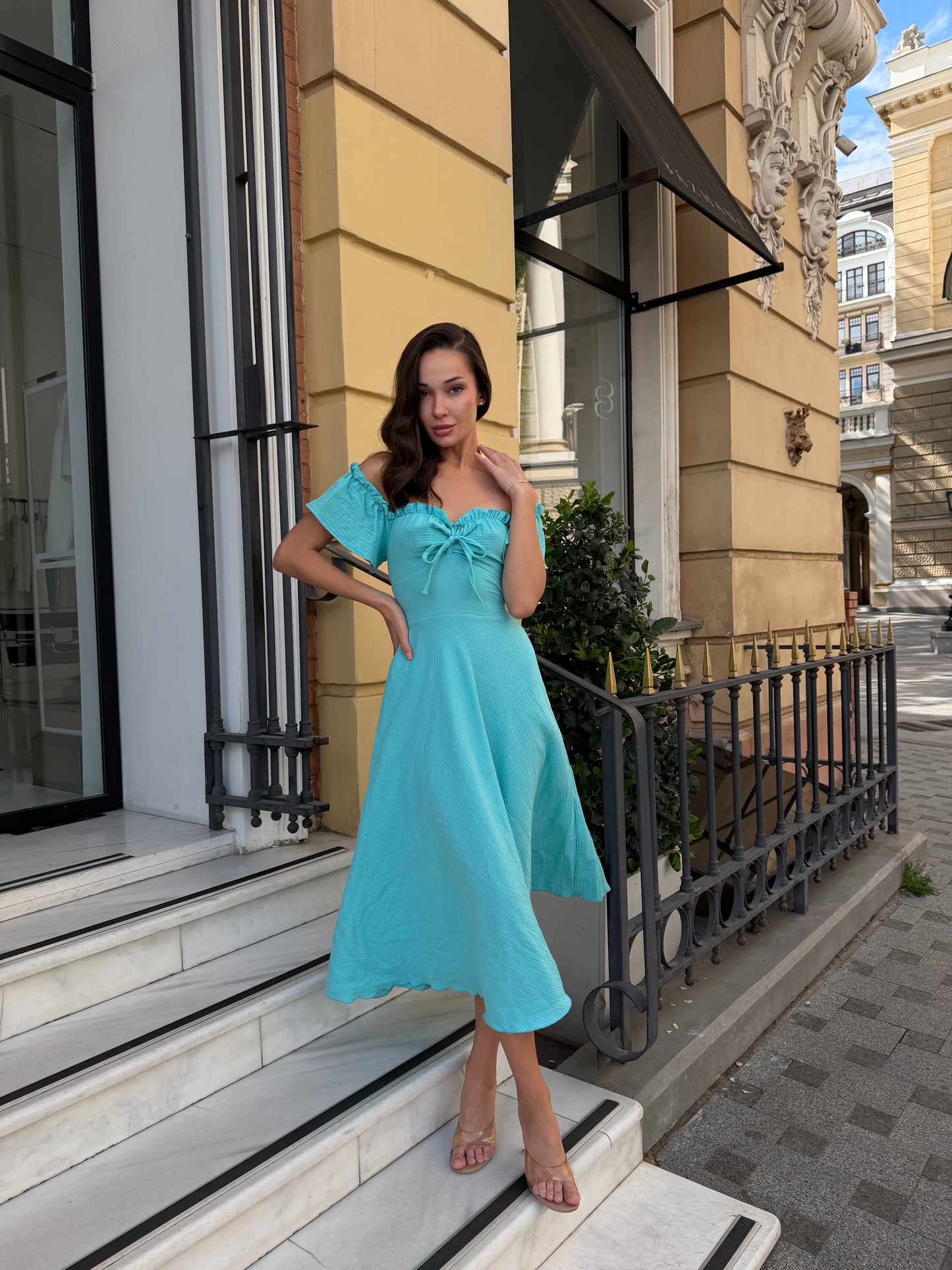 A woman wearing a light turquoise off-the-shoulder midi dress with a sweetheart neckline and a bow detail at the bust, standing on white marble steps in front of a beige building with black railings.