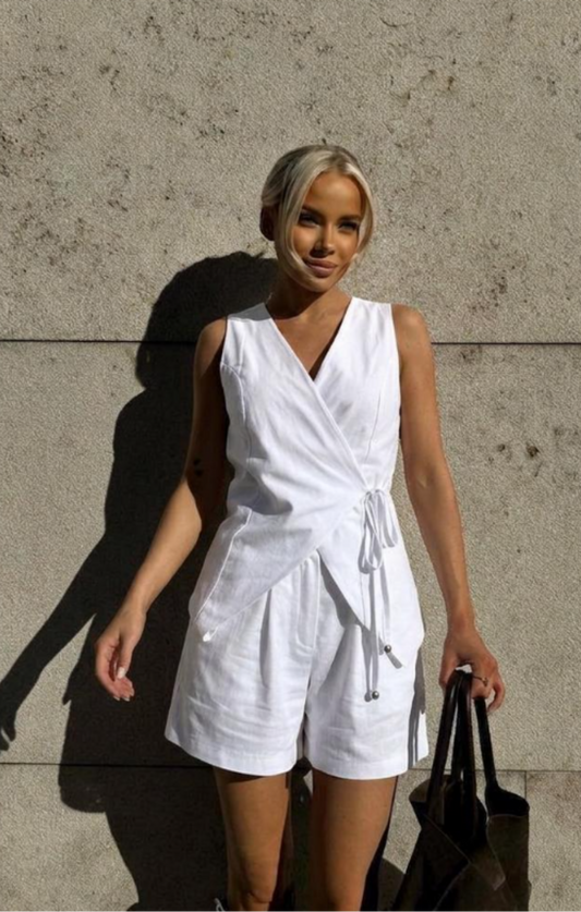 A woman wearing a white sleeveless vest linen matching set shots with shorts stands against a textured concrete wall, holding a black tote bag.